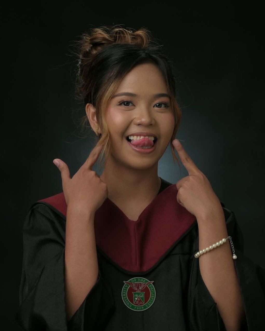 Sharms in a University of the Philippines graduation gown, grinning with tongue out and pointing at the grin with both index fingers.